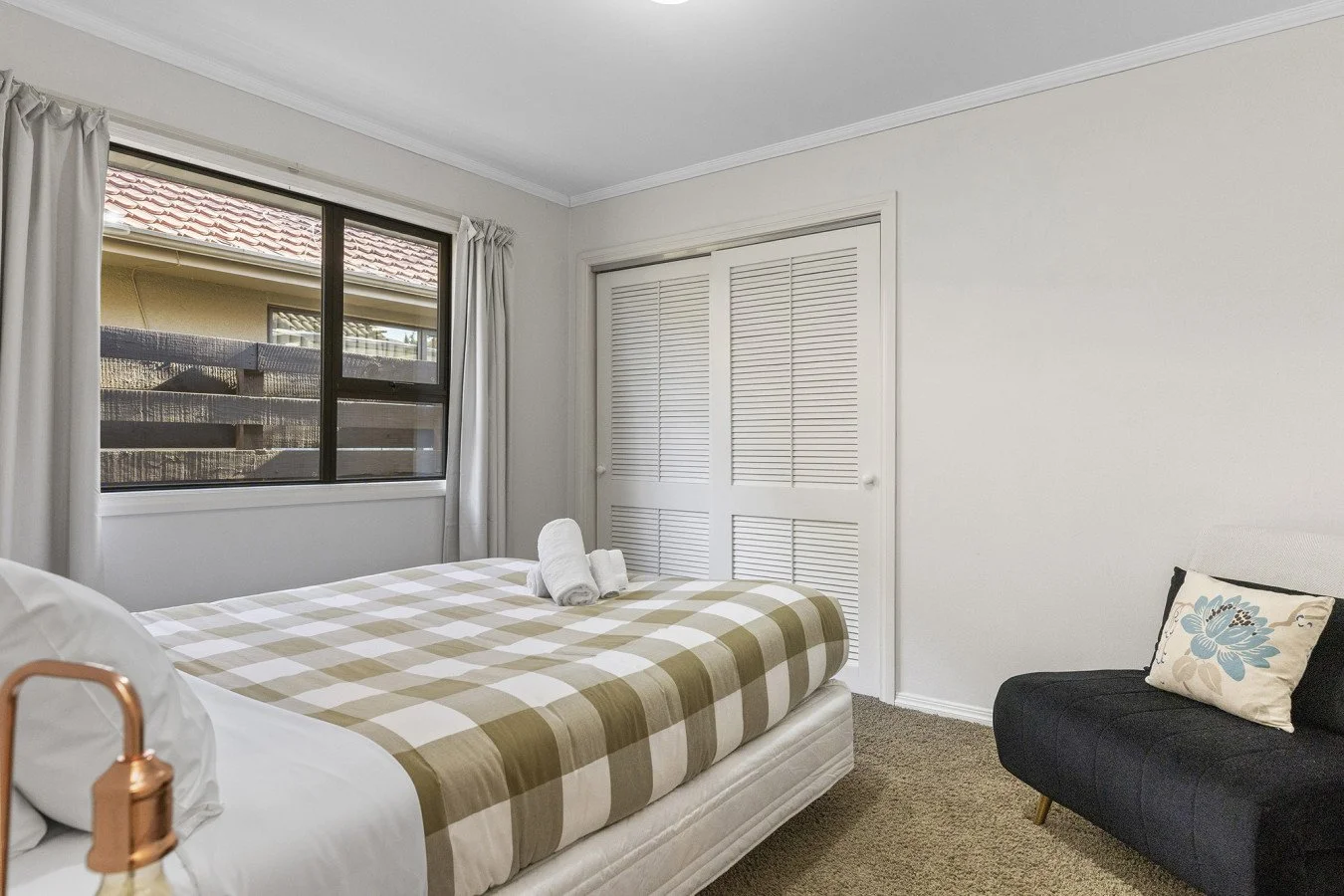 A bedroom with a bed that has a checkered beige and white bedspread, two rolled towels on the bed, a black sofa with a floral pillow, a window with curtains, and a closet with louvered doors.