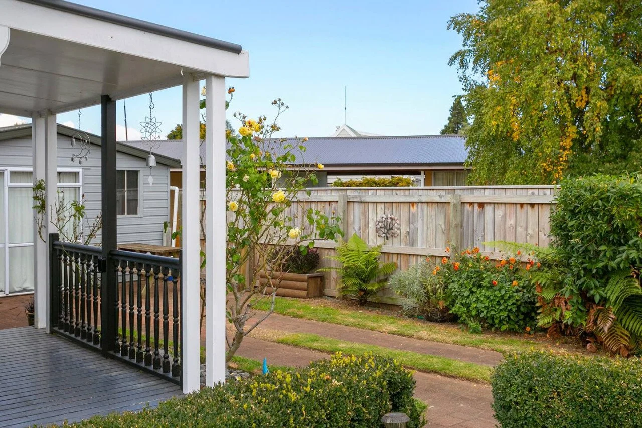 A backyard garden with a wooden porch on the left, a wooden fence in the background, and various green plants and flowers, including yellow roses and orange blossoms, on a sunny day.