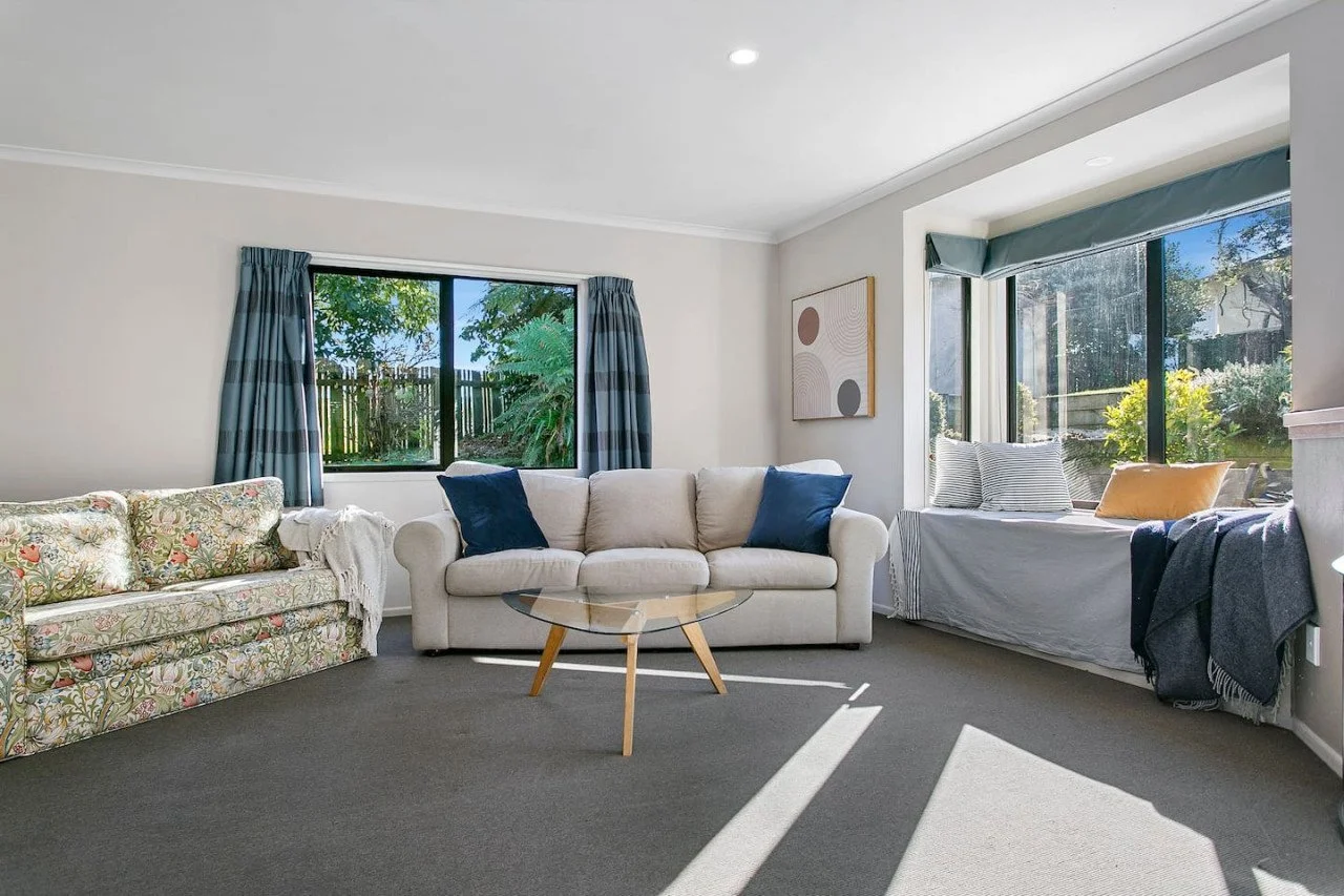 Bright living room with three windows showing outdoor greenery, a floral sofa on the left, a beige sofa with blue pillows in the middle, a window seat with pillows on the right, a modern abstract wall art, and sunlight casting shadows on a gray carpe