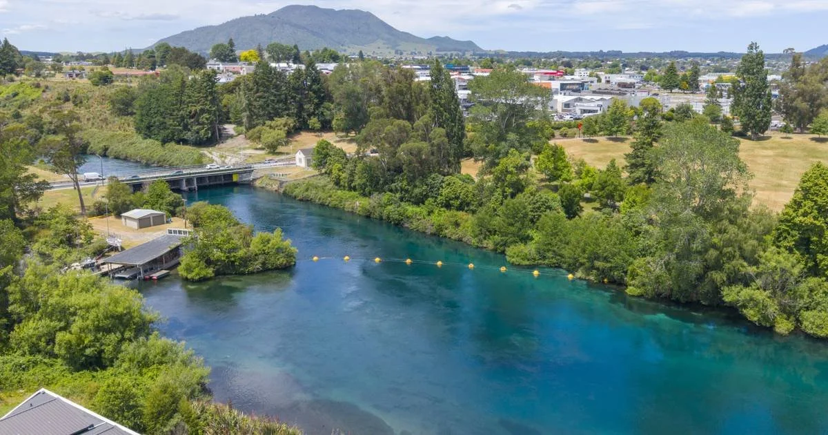 A scenic view of a river with clear blue water flowing under a bridge, surrounded by green trees, with a town or industrial area in the background and mountains in the distance.