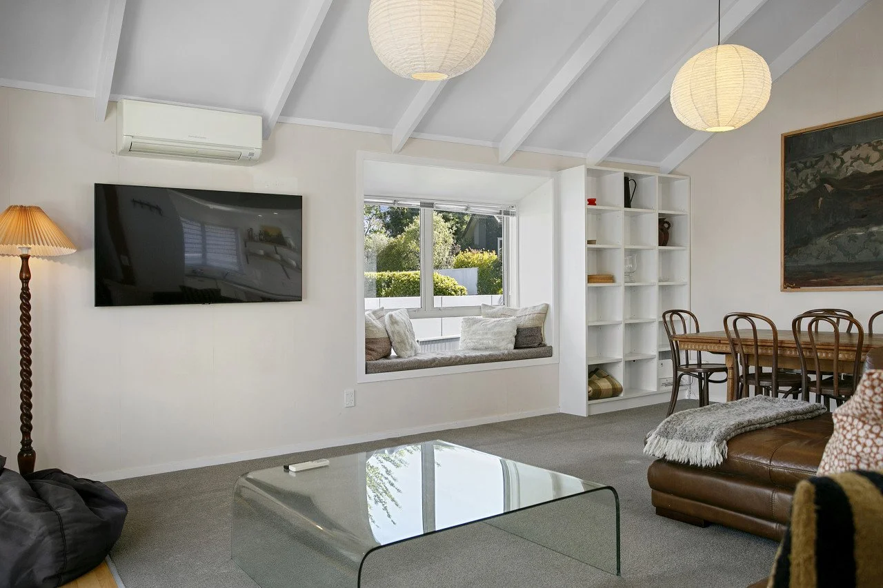 Living room with a wall-mounted flat-screen TV, a window seat with cushions, a built-in white bookshelf, a wooden dining table with chairs, a leather sofa with a blanket, a floor lamp, and two hanging paper lanterns.