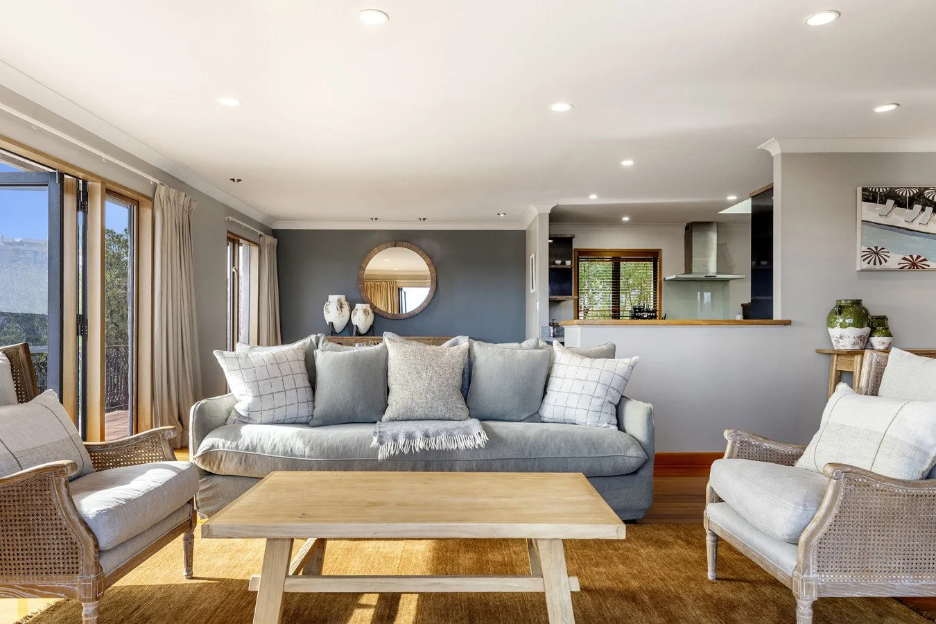 Living room with large sofa, armchairs, wooden coffee table, and sliding glass doors allowing natural light, decorated in neutral tones with curtains and decorative vases.