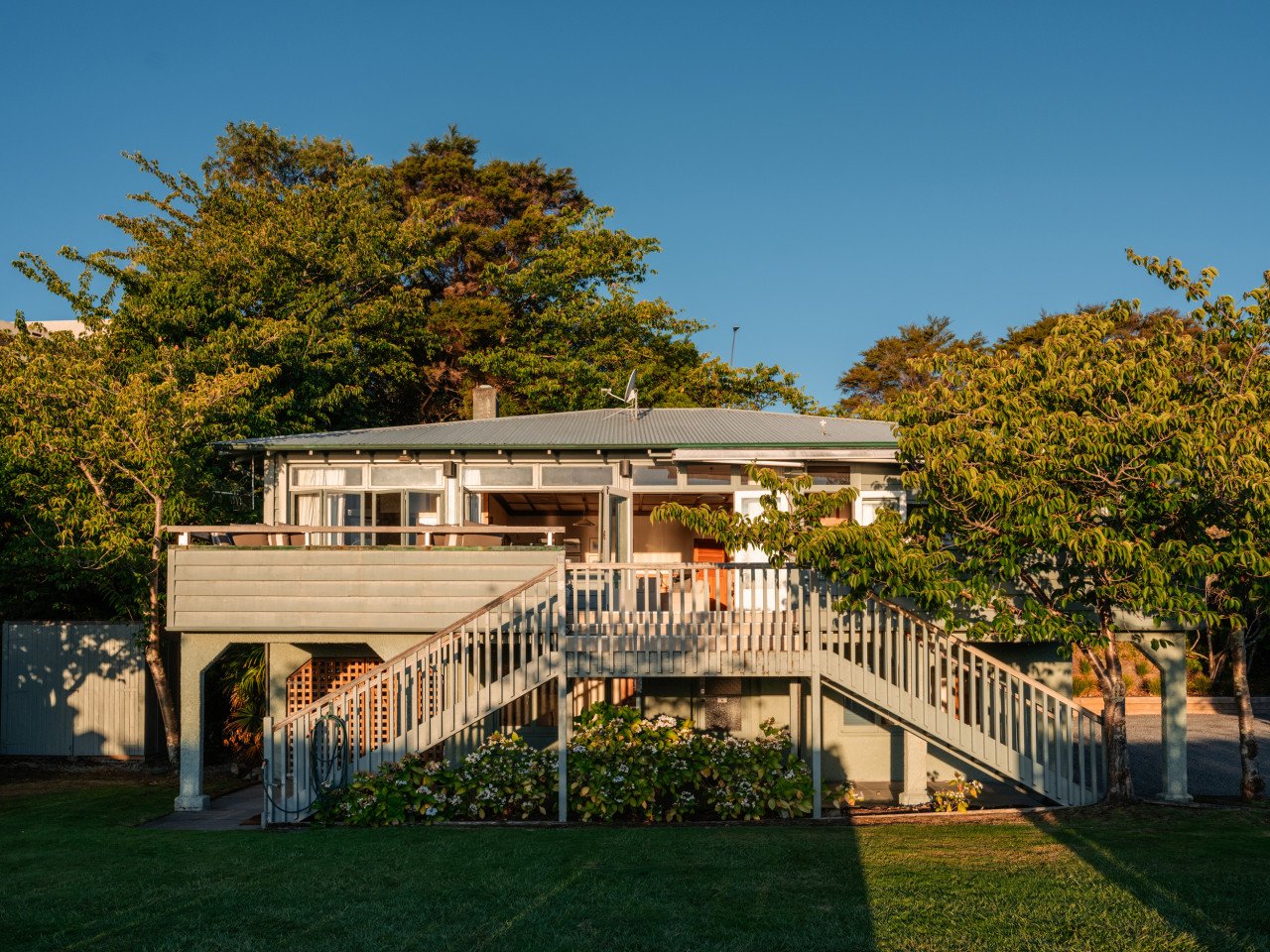 Back of a house elevated on stilts with a staircase leading up to a balcony, surrounded by trees and green lawn.