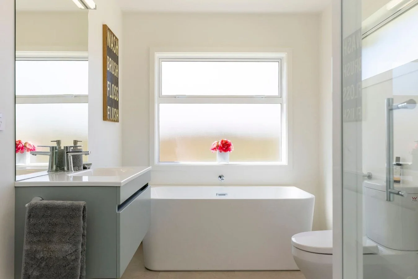 Modern bathroom with a bathtub under a large window, a vanity with a sink, a towel, a flower vase, and a toilet.