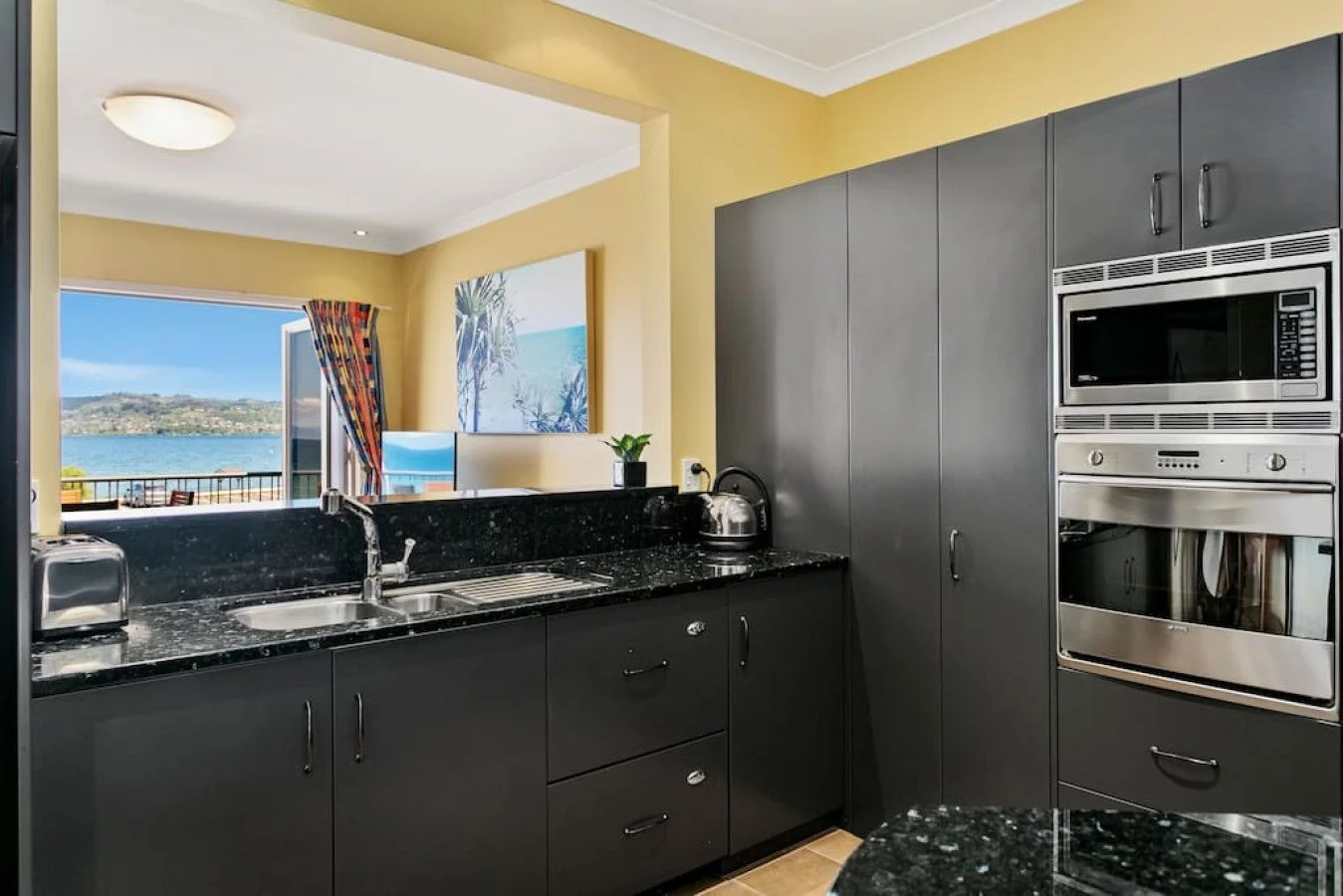 Modern kitchen with black countertops, gray cabinets, stainless steel appliances, a window showing a view of water and hills, and yellow walls.
