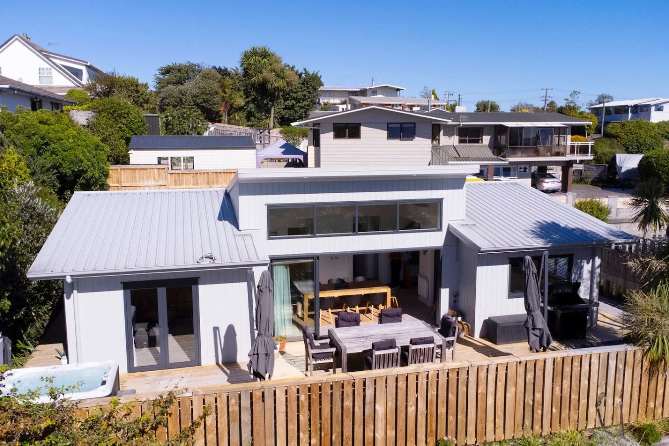 A modern house with a white exterior and metal roof, featuring an outdoor deck with a dining table, umbrellas, and a hot tub, enclosed by a wooden fence, located in a hilly neighborhood with other houses and lush greenery.