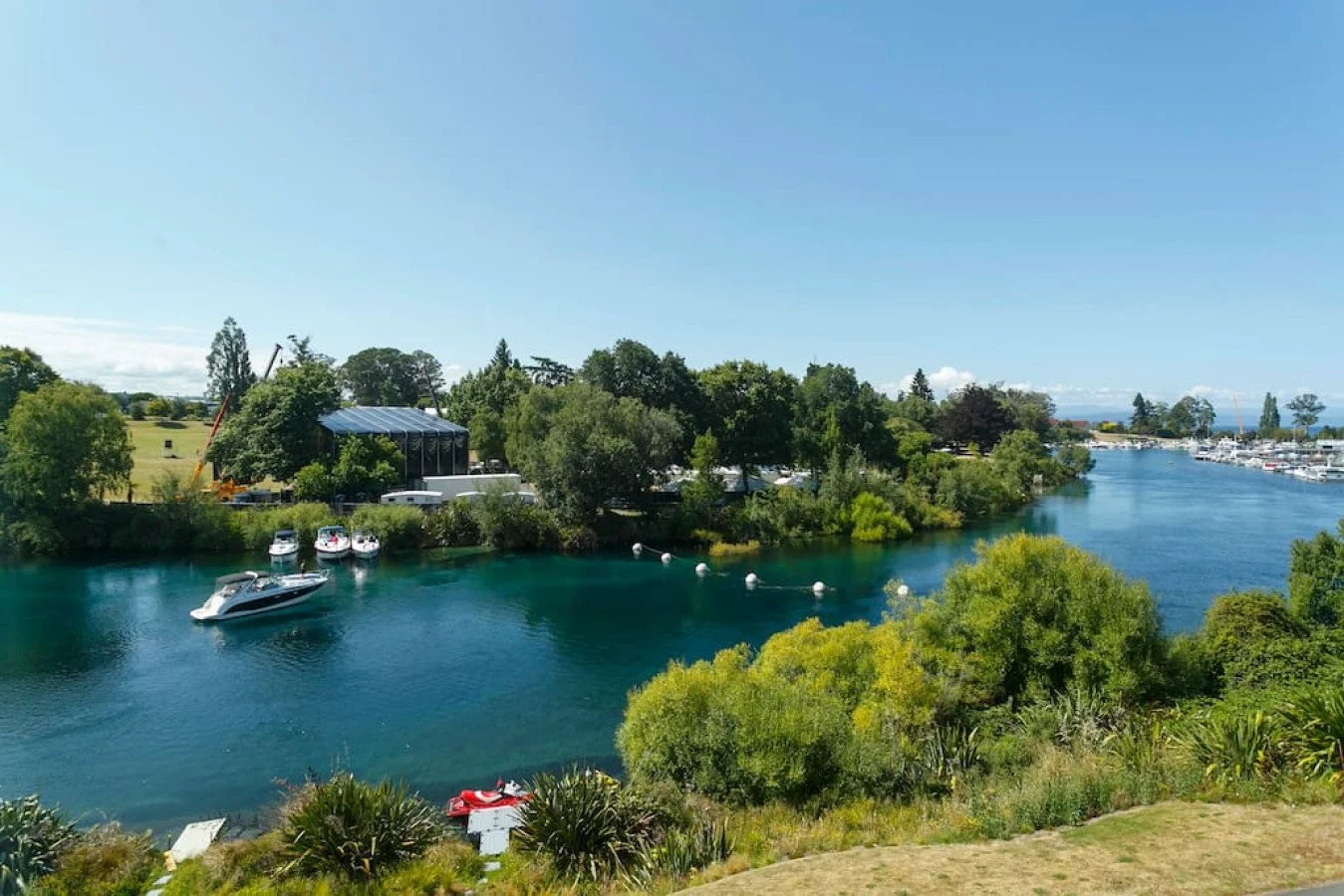 Scenic view of a marina with boats docked in blue water, surrounded by green trees and vegetation, under a clear blue sky.