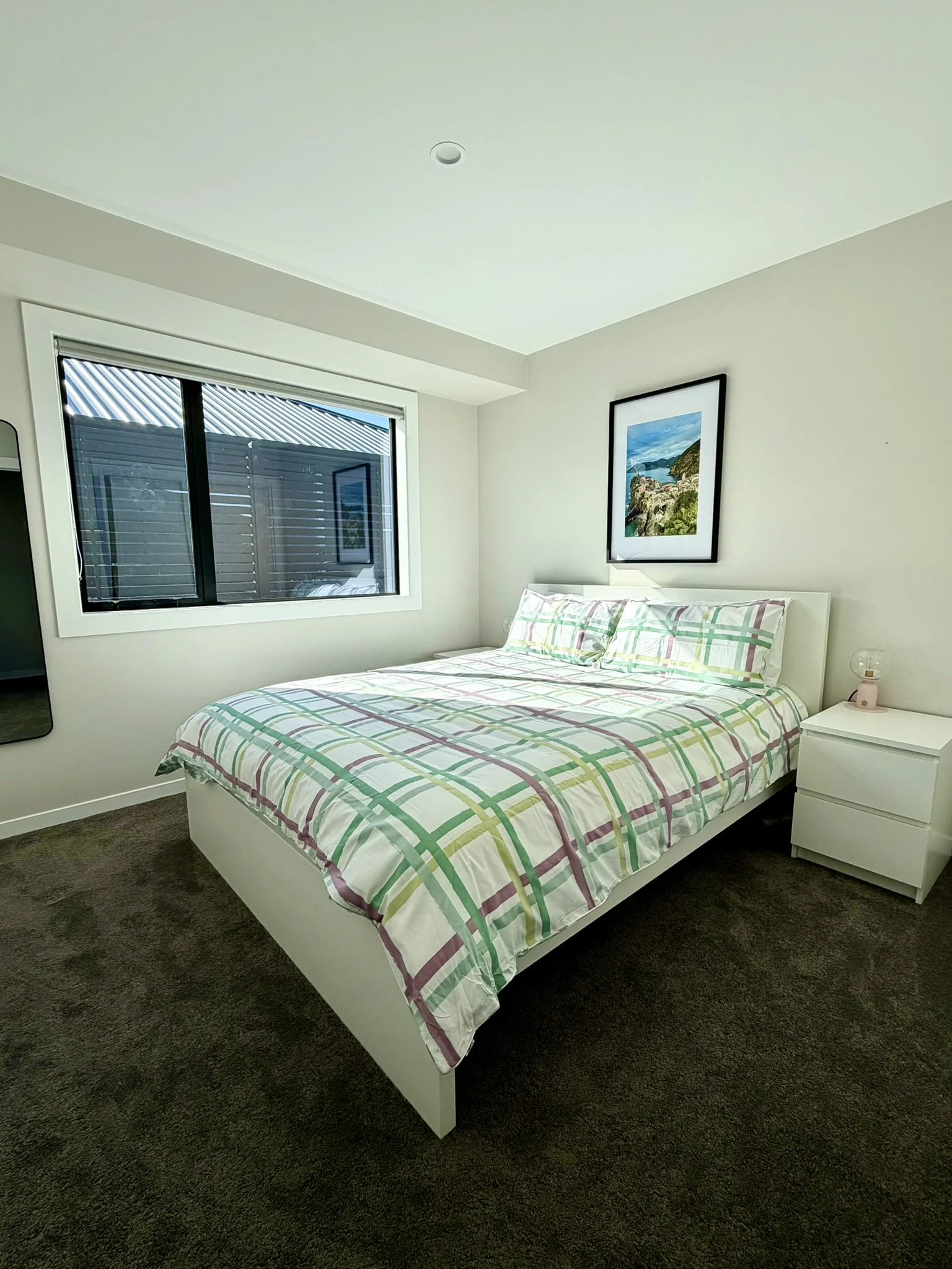 A bedroom with a white bed and colorful plaid bedding, a white nightstand with a lamp, a large window, and a framed landscape photo on the wall.
