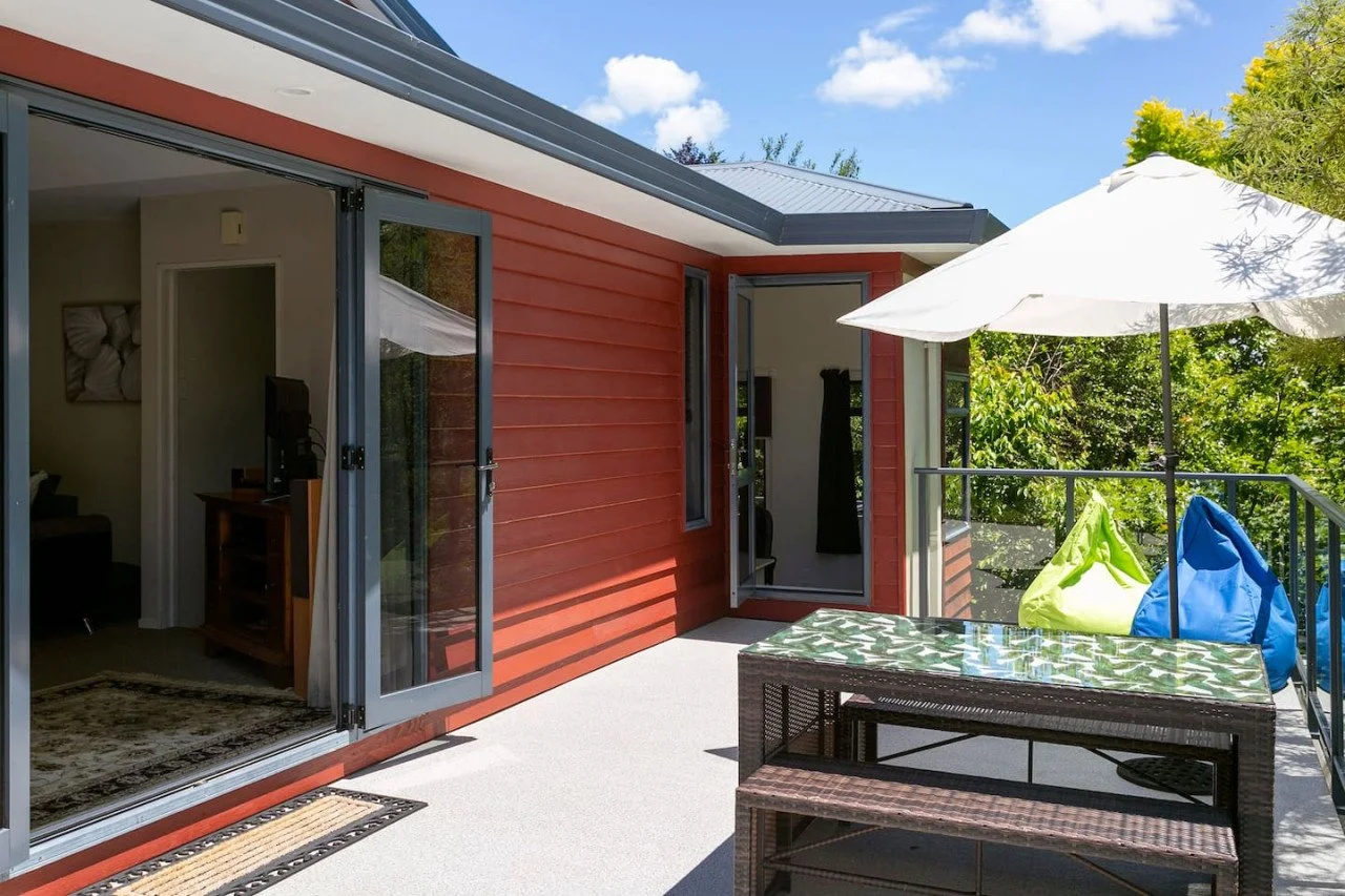 Outdoor balcony with glass door leading inside a house, umbrella, colorful bean bags, and lush green trees in the background.