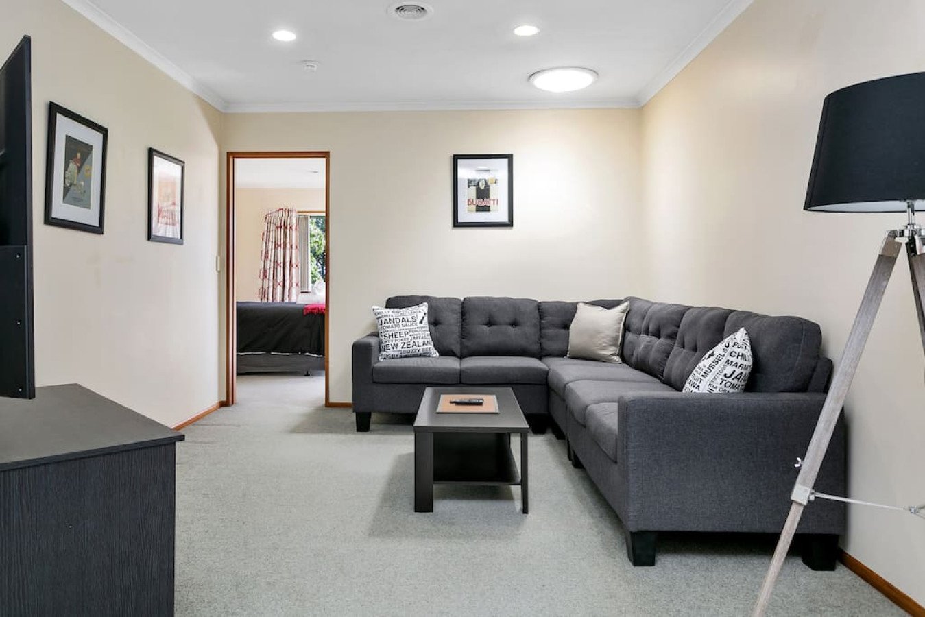 Living room with gray sectional sofa, black coffee table, and a stand-up lamp with black shade. The walls are beige, decorated with framed art, and a doorway leads to a bedroom with a window and patterned curtains.