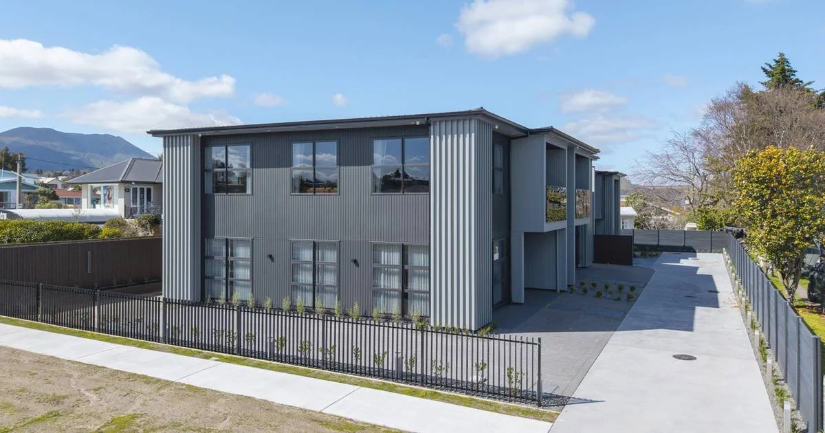 Modern two-story residential building with metal siding, large windows, and a driveway, surrounded by a black fence and trees, with mountains in the background.