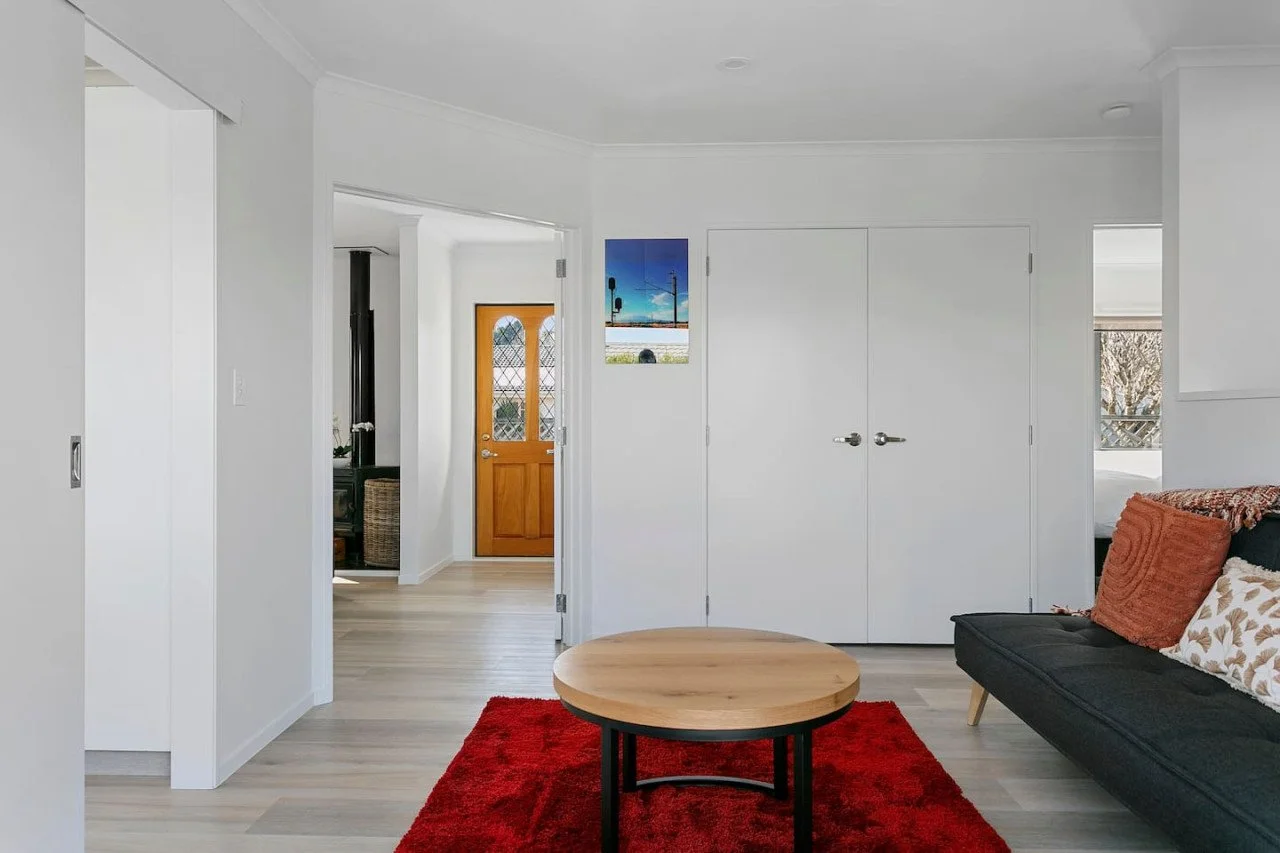 Living room with a black sofa, orange and patterned throw pillows, a round wooden coffee table, a red rug, white walls, and a door leading to the outside with a wooden door and a window.