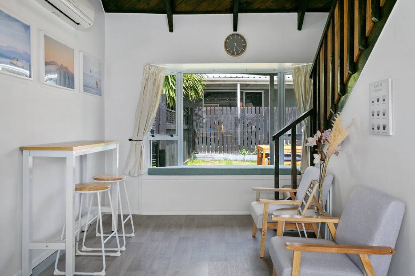 Living room with large front window, white walls, wooden ceiling beams, chairs, wall art, clock, and outdoor view with garden