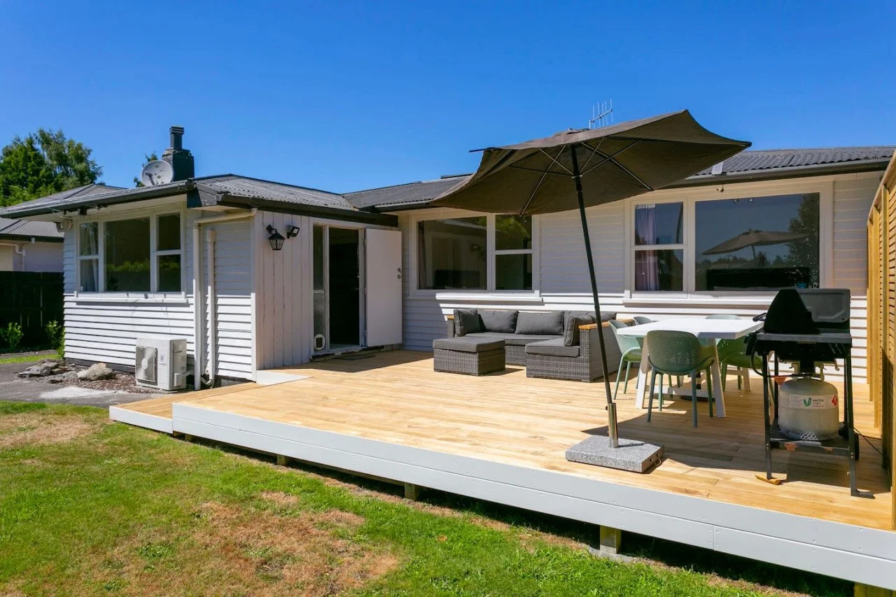 Backyard patio with outdoor furniture, umbrella, dining table, barbecue grill, and house with white siding and large windows.