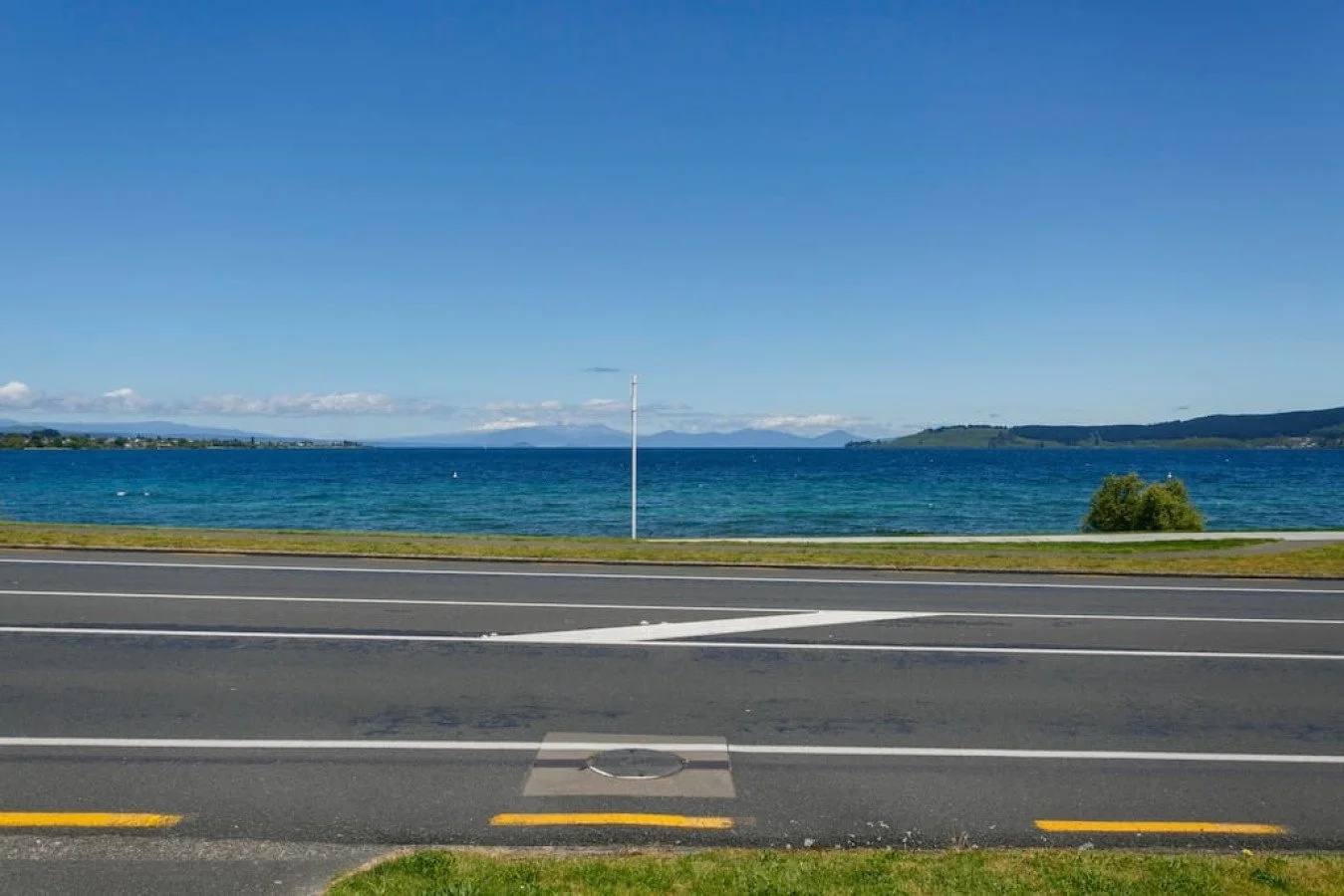 View of a shoreline with a body of water, distant land, and a clear blue sky, taken from the roadside with visible road markings in the foreground.