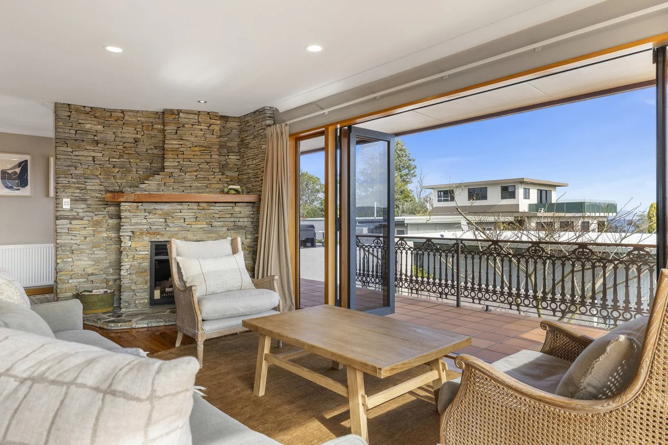 Living room with stone fireplace, beige armchairs, wooden coffee table, and open balcony doors showing a clear sky and neighboring buildings.
