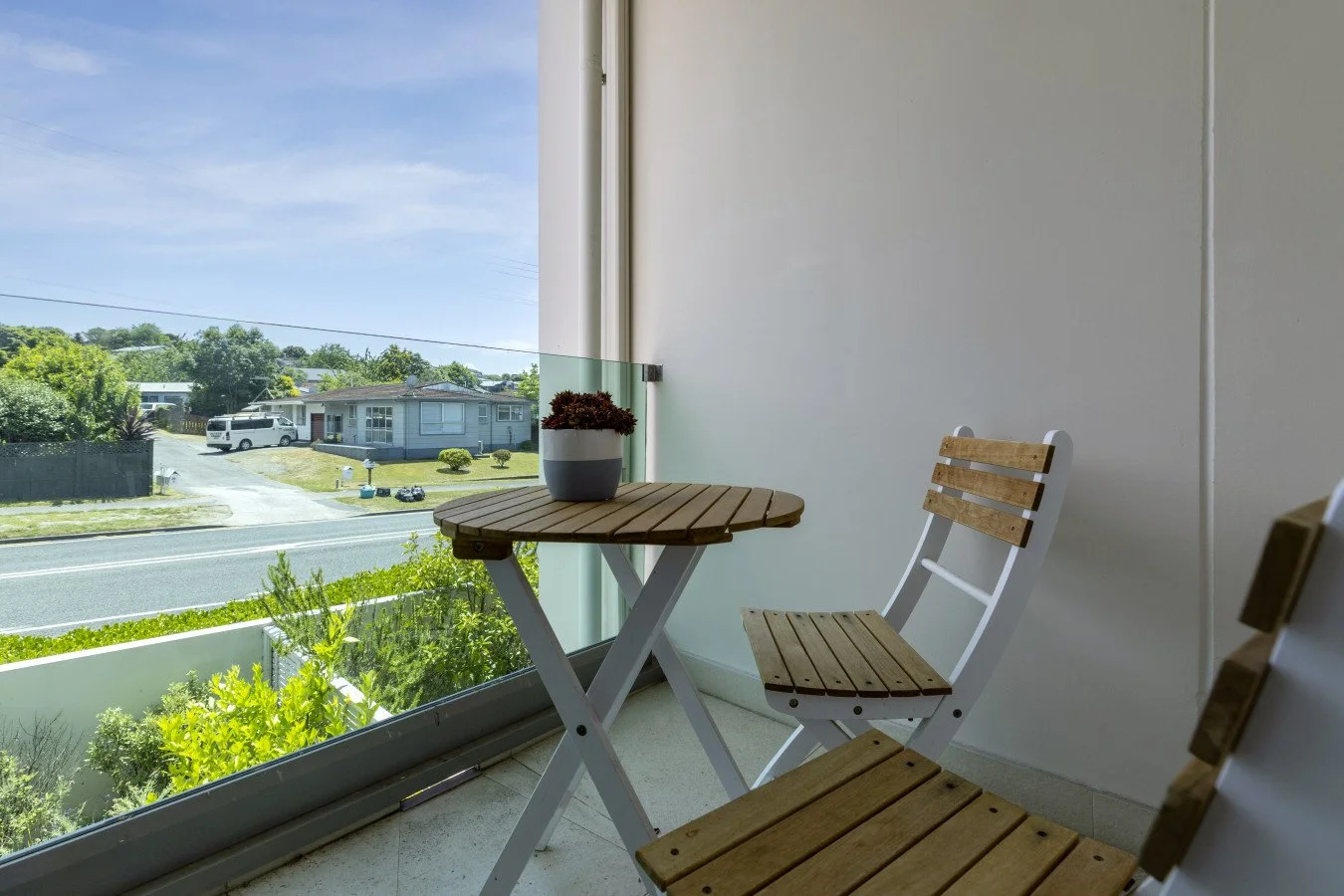 Small balcony with a wooden table and two wooden chairs, a planter with a plant, and a view of suburban houses and greenery outside.