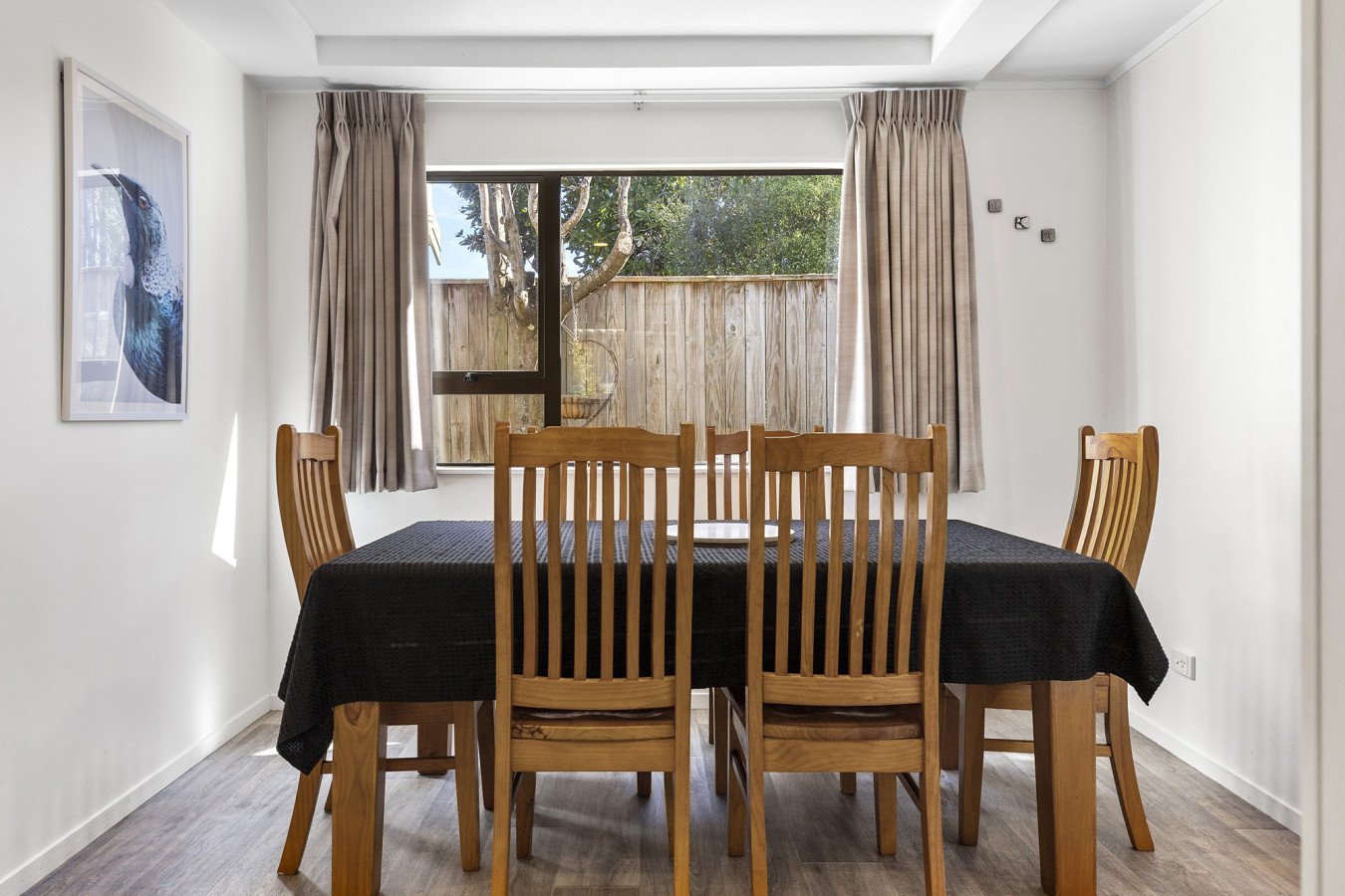 Dining room with wooden table, black tablecloth, six wooden chairs, window with beige curtains, outside view of trees and fence, framed art of bird on white wall.