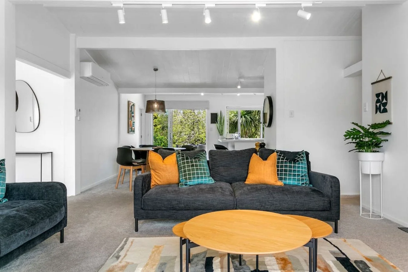 Living room and dining area with black couch, yellow and green pillows, wooden coffee table, dining table with black chairs, large window with greenery outside, white walls, and modern decor.