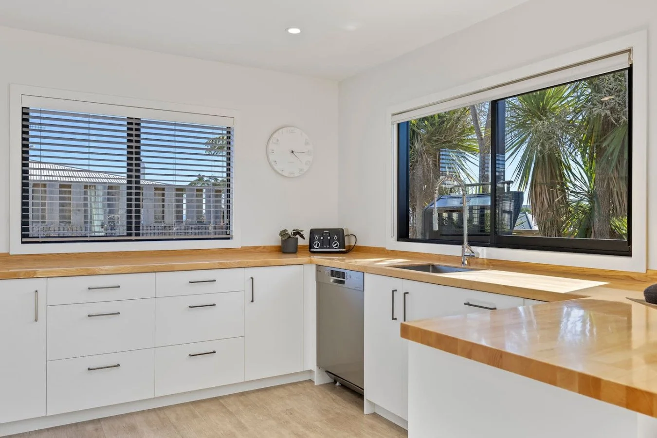 Bright kitchen with white cabinets, wooden countertop, black window frames, and large windows showing trees outside.