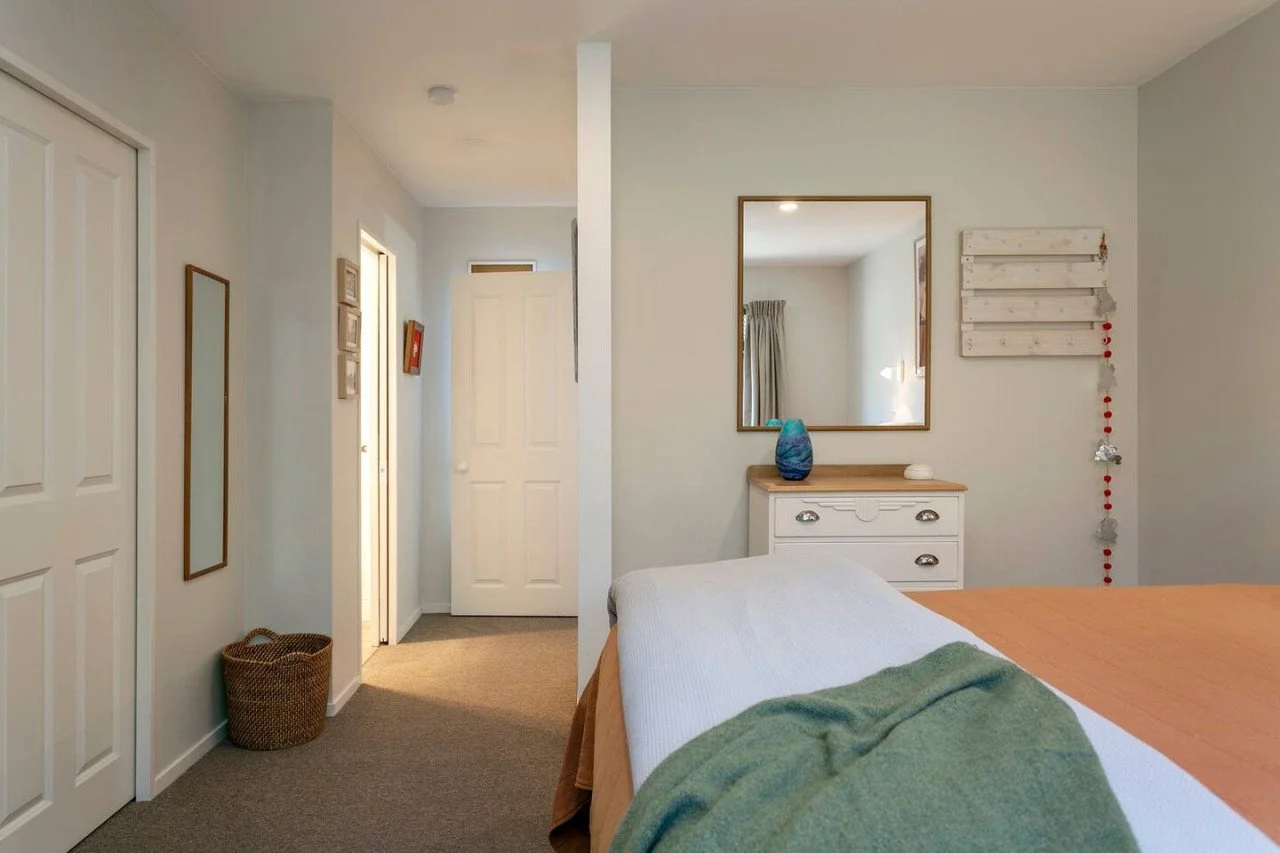 A cozy bedroom with light-colored walls, a bed with white and orange bedding topped with a green blanket, a dresser with a mirror above it, and a window with curtains.