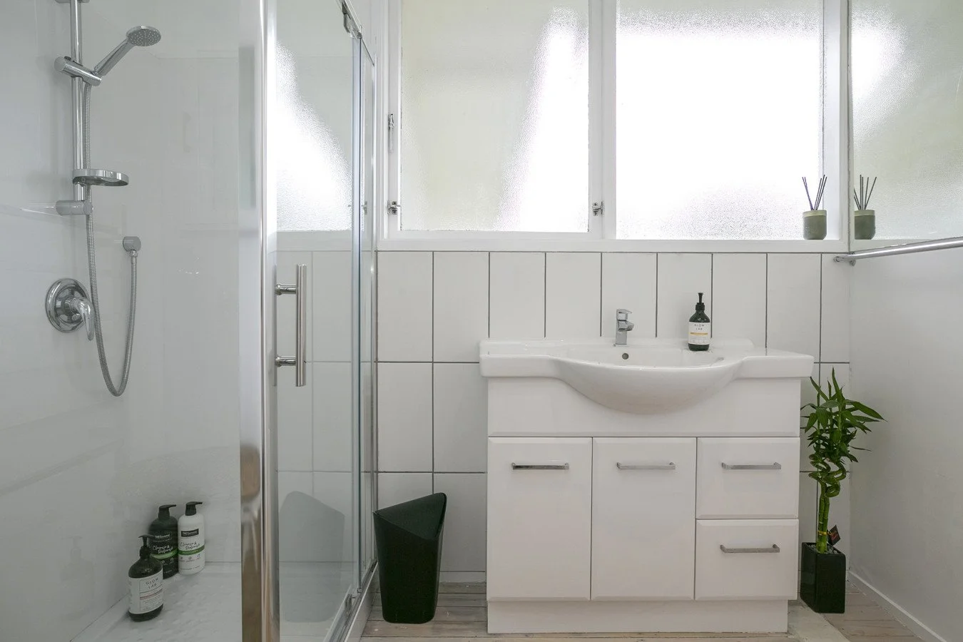 Bathroom with white tiles, a frosted window, a glass shower enclosure on the left, and a white vanity with a sink and cabinet drawers. There are potted plants and toiletries on the countertop and a small plant in a black pot on the floor.