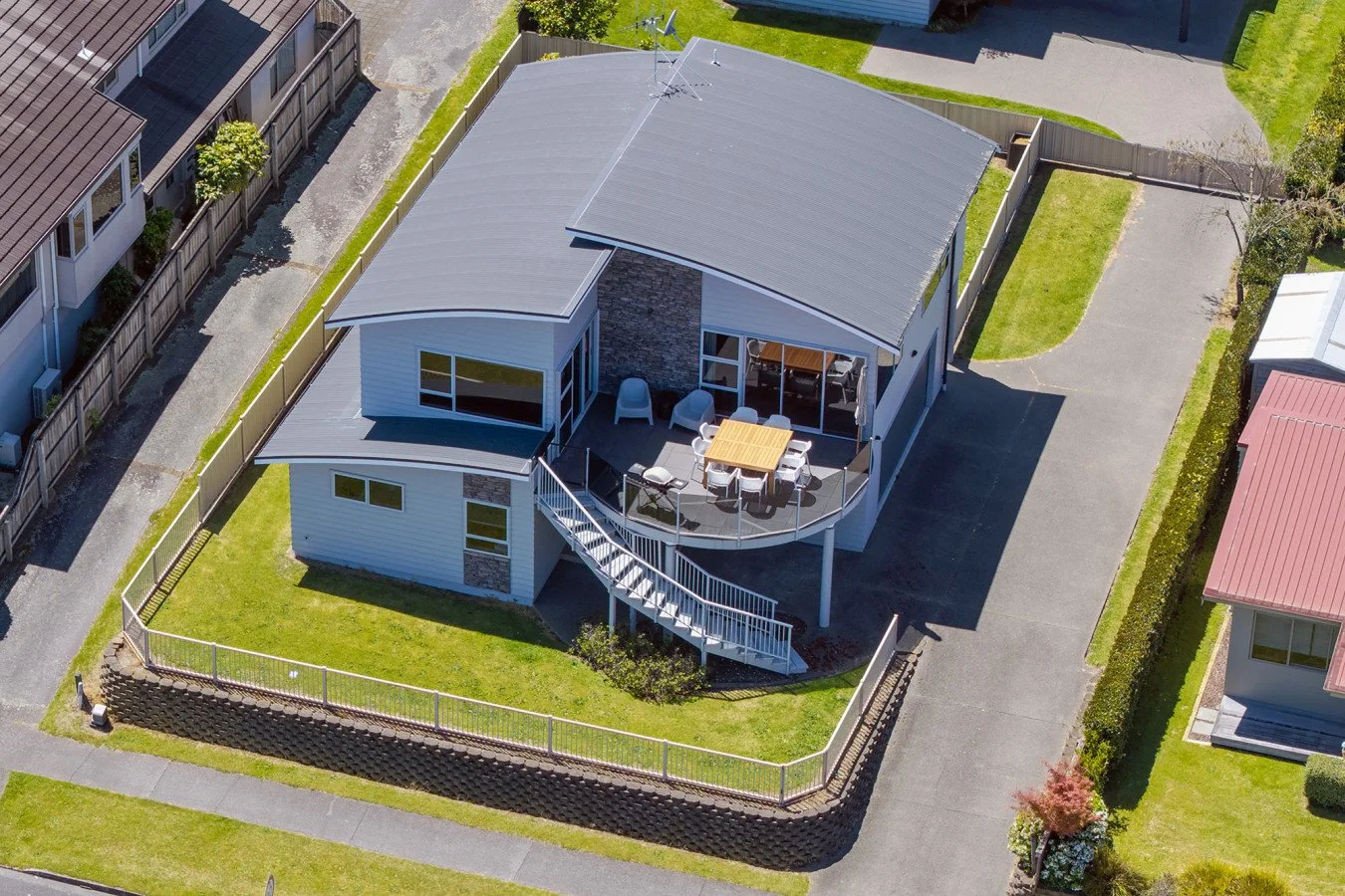 An aerial view of a modern house with a curved roof, a deck with outdoor furniture, and a driveway. The house is surrounded by a lawn and neighboring houses.