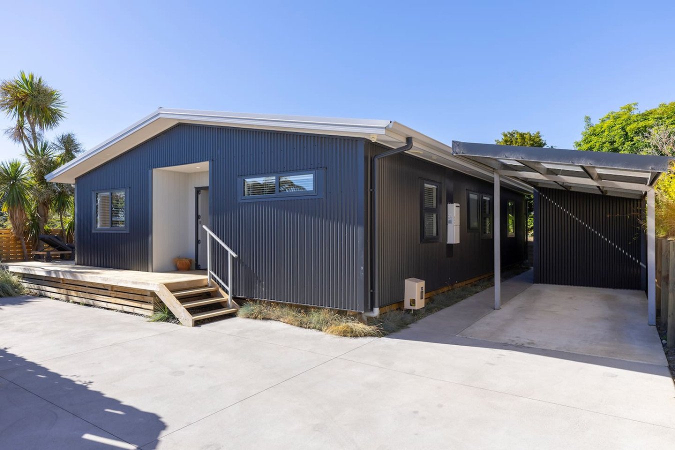 Modern single-story house with dark exterior walls, small front porch, and an attached carport on the side, with a concrete driveway and some desert plants nearby.