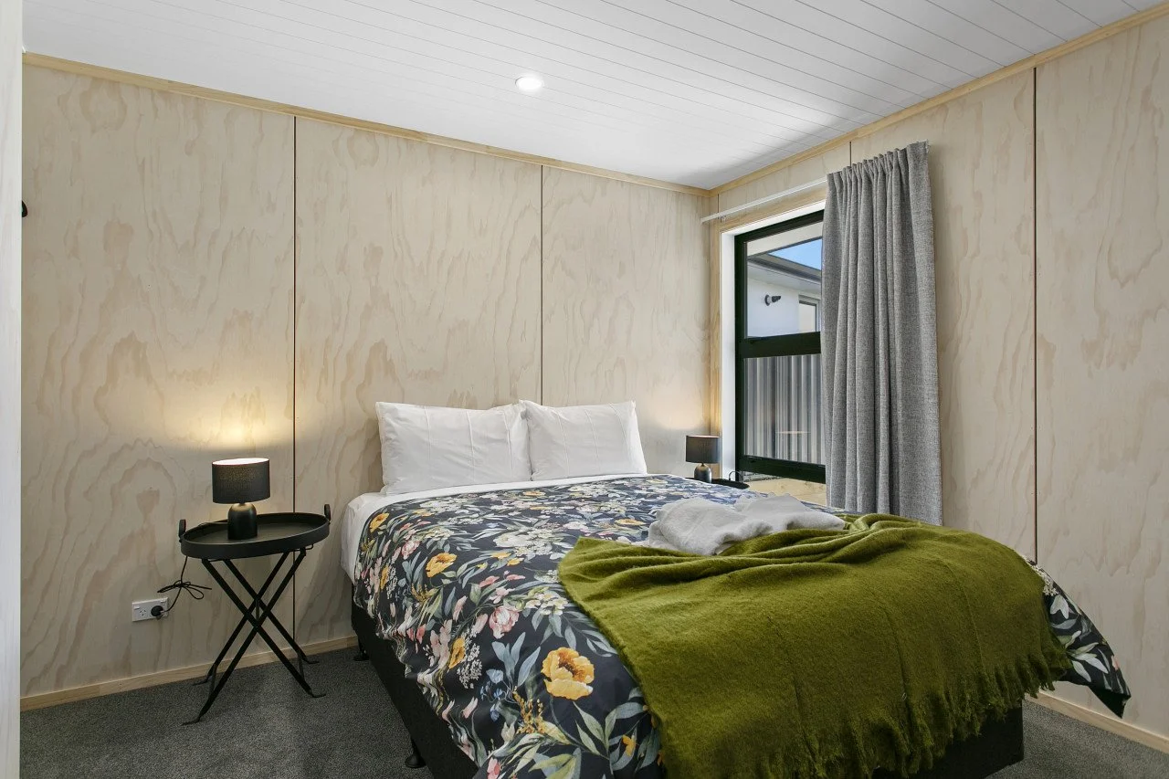 A cozy bedroom with a bed covered in a floral duvet, a green throw blanket, and white pillows. There are two black bedside tables with small black lamps on each side, and a window with gray curtains letting in natural light.