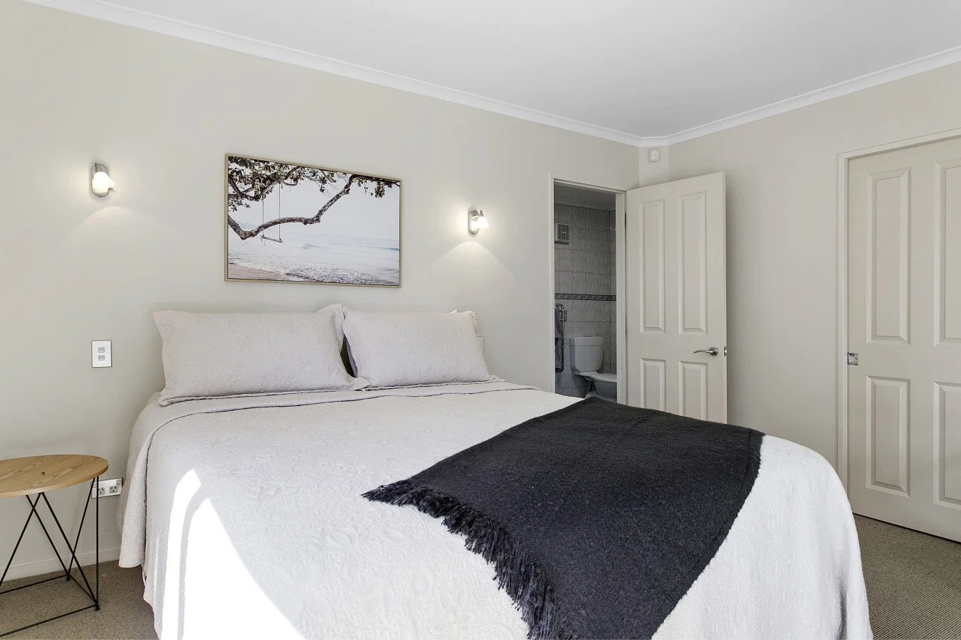 Bedroom with white bedding, black throw blanket, wall-mounted lights, framed seaside artwork, bedside table, open door leading to a bathroom with toilet.