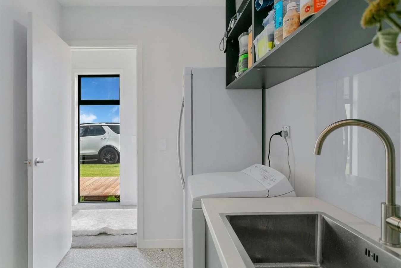 Laundry room with a white top-loading washing machine, white refrigerator, black shelving with cleaning supplies, stainless steel sink with a faucet, white wall, and a door with a large window leading outside to a car parked on grass.