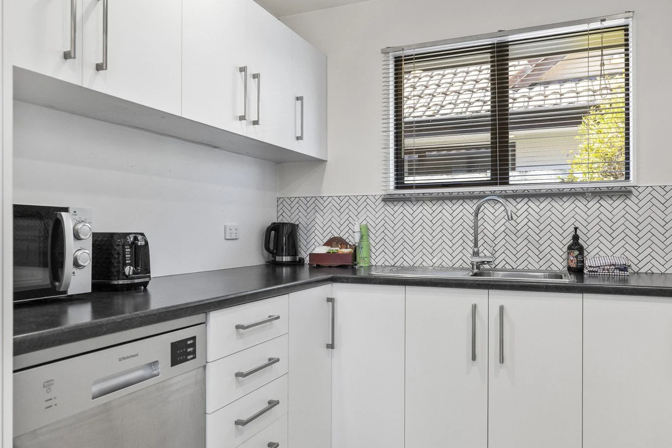 Modern kitchen with white cabinets, black countertops, stainless steel appliances, and a window with blinds over the sink.