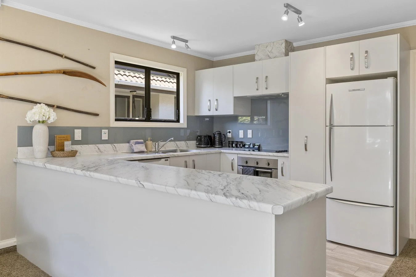Modern kitchen with white cabinets, marble countertops, and a white refrigerator, with decorative antique tools on the wall near a window.