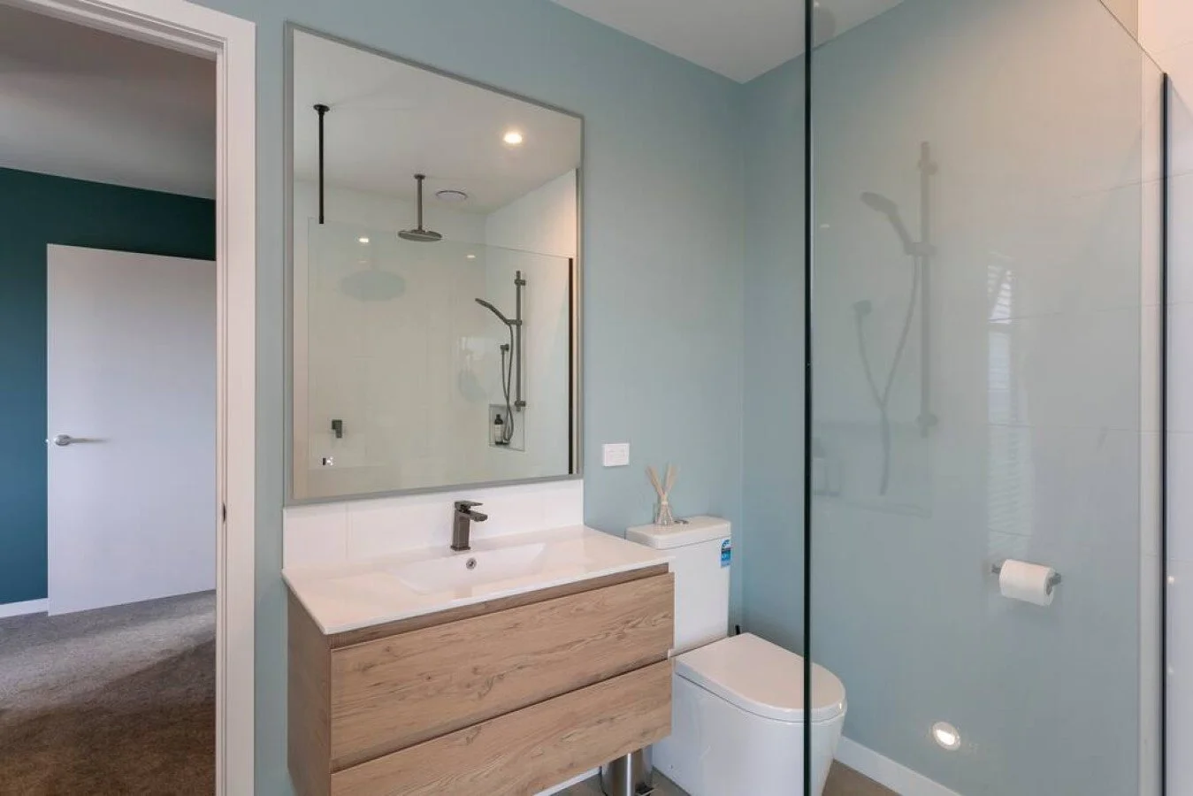 Modern bathroom with a wooden vanity, large mirror, glass shower enclosure, and toilet, with a teal accent wall and beige carpeted floor.