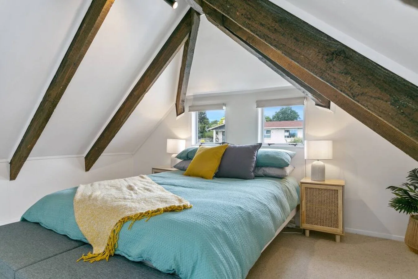 Bedroom with sloped ceiling, bed with teal bedding and pillows, two windows, nightstands with lamps, and a small plant in the corner.