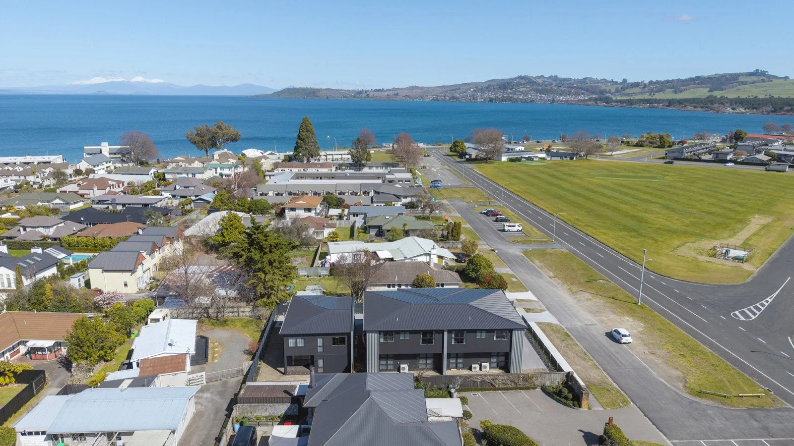 Aerial view of residential houses near a body of water with mountains in the background, green open field, and roads in a suburban area.