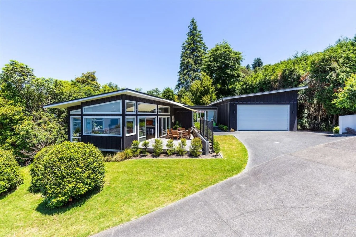 Modern black house with large glass windows, outdoor patio with a table and chairs, surrounded by green grass, bushes, and trees, on a sunny day.