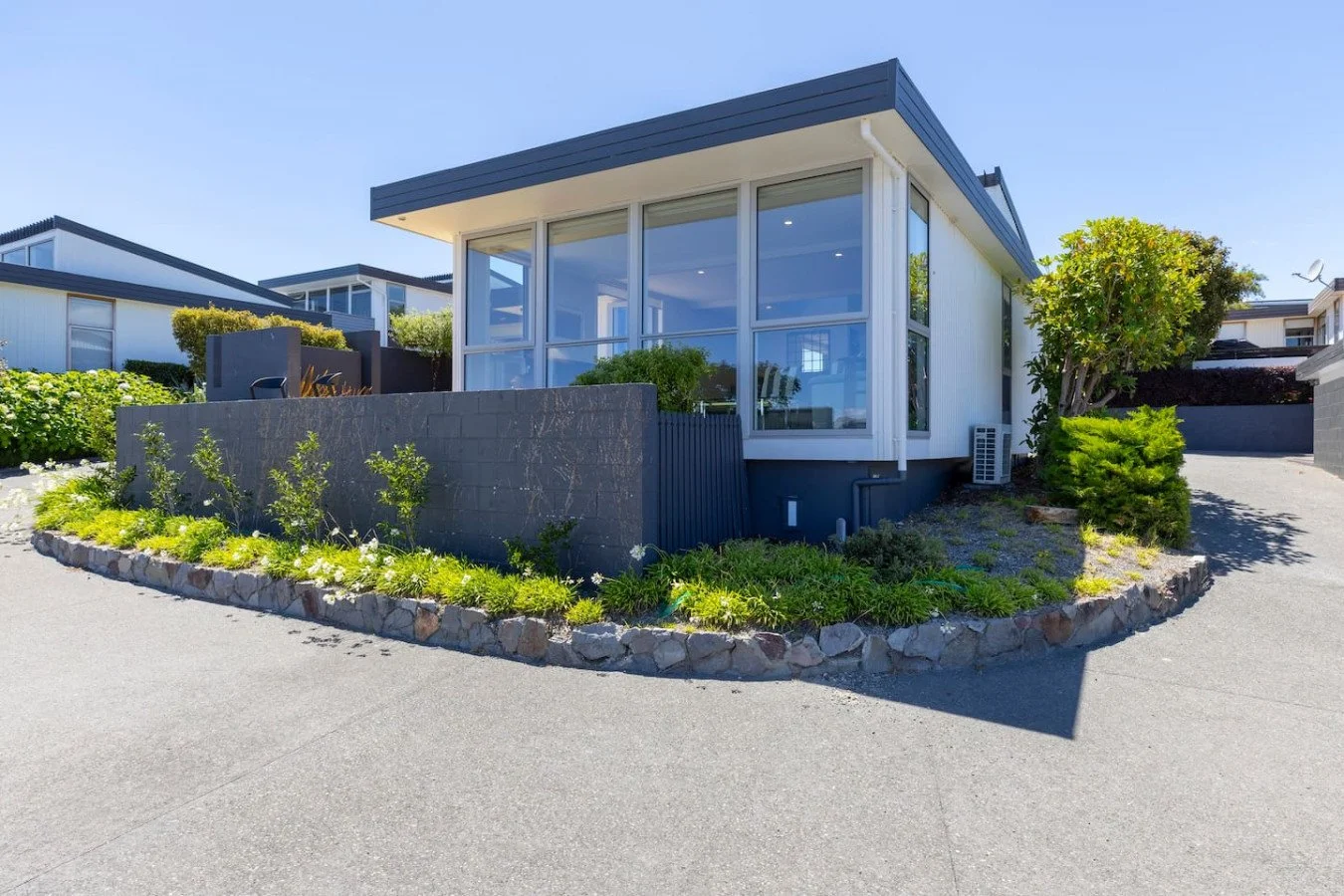 Modern house with large windows, landscaped front yard with green plants, trees, and a stone border, under a clear blue sky.