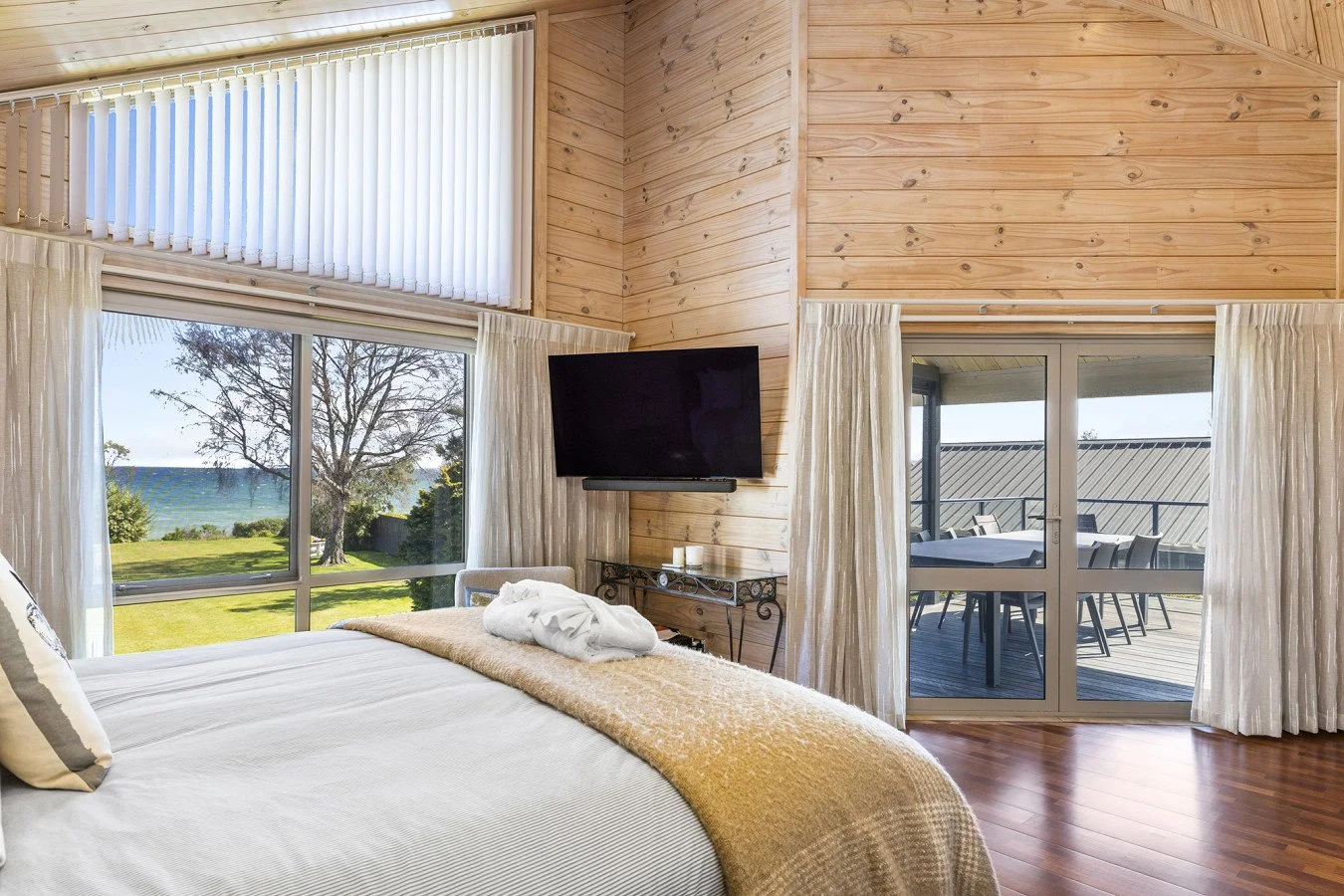 A bedroom with large windows overlooking a grassy yard and ocean. The room has light wood walls, a mounted flat-screen TV, and a sliding glass door leading to an outdoor patio with a table and chairs.