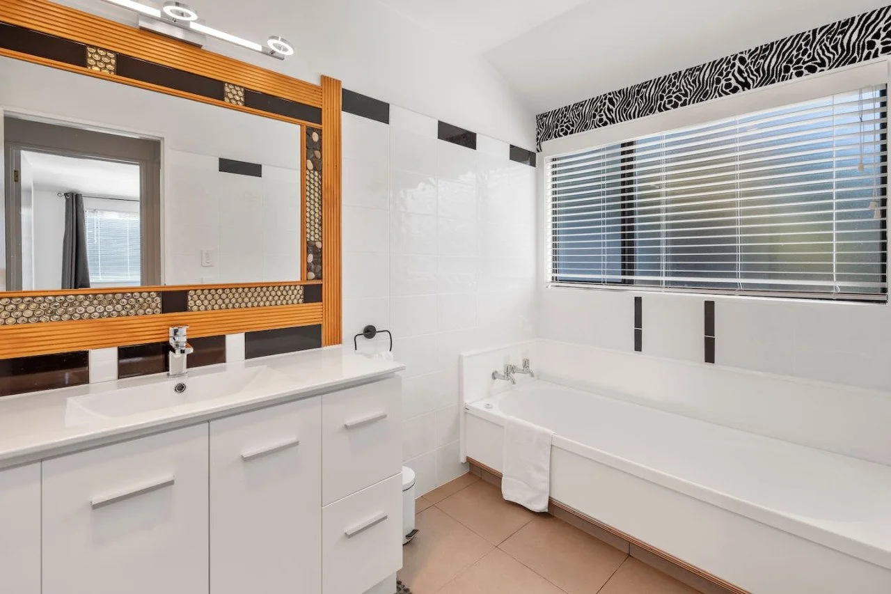 Bathroom with a large mirror, white vanity with a sink, bathtub, and window with blinds.