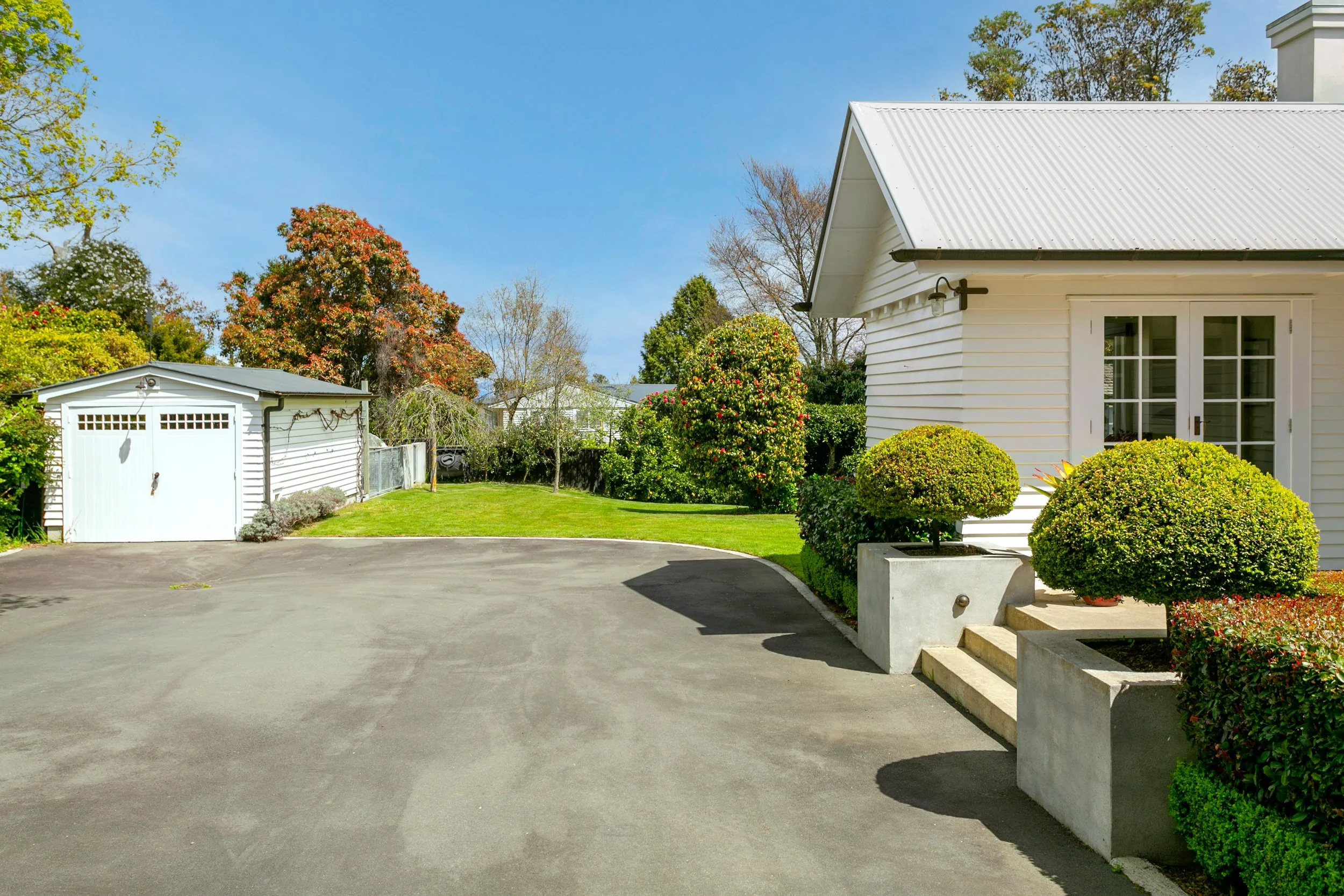 A suburban backyard with a white house, trimmed bushes, a green lawn, trees, and a white shed on a clear day.