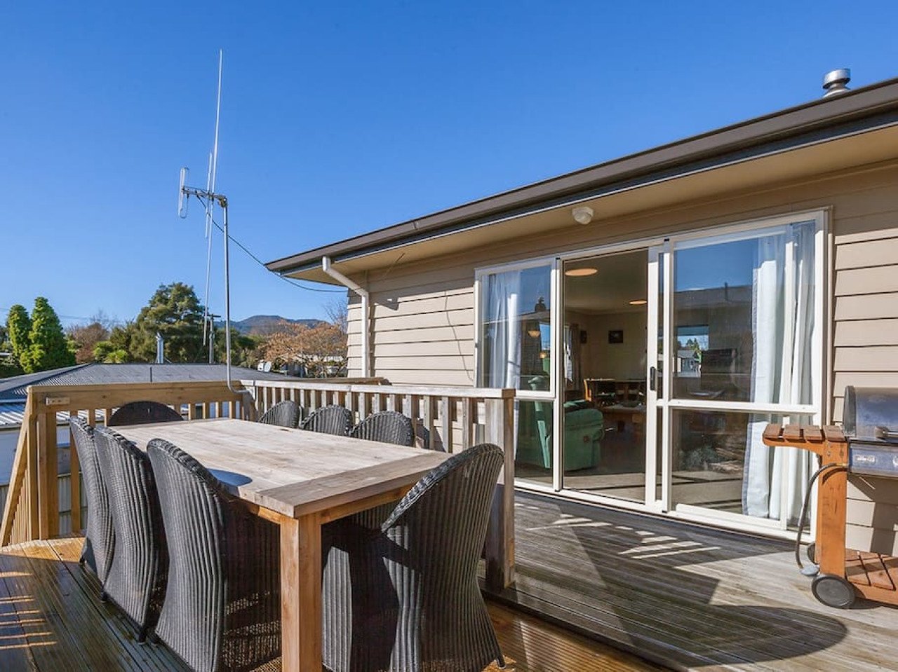 Deck with outdoor dining table and chairs, attached to house with sliding glass door, grill on side, and clear blue sky overhead.