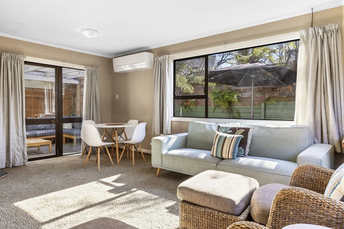 Living room with beige walls, a large window, a light-colored sofa, wicker chair, round dining table with chairs, and sliding glass door leading to an outdoor patio with an umbrella.