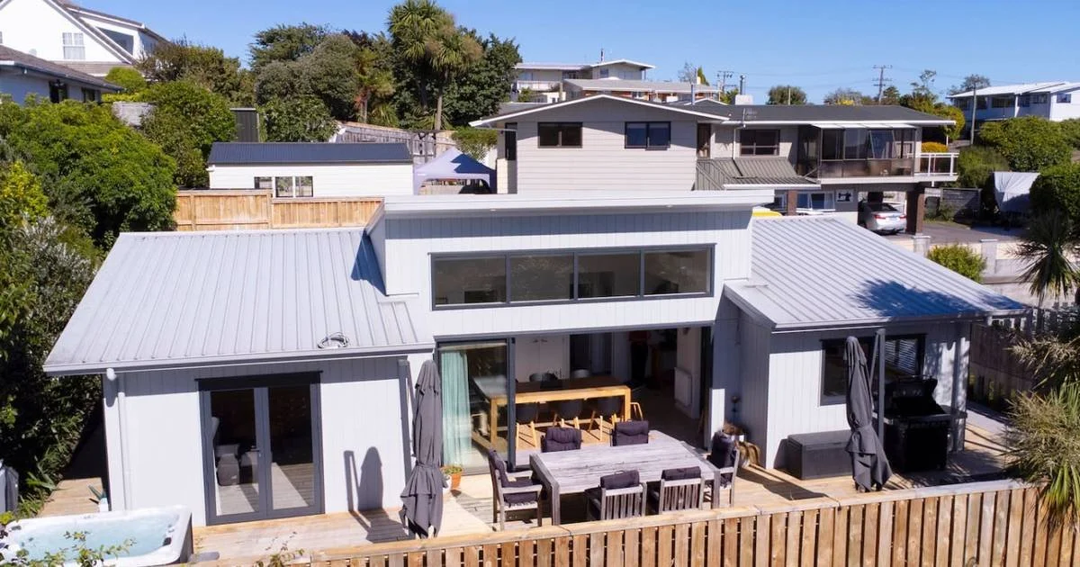 Modern house with gray exterior, metal roof, and outdoor patio featuring dining table, chairs, and umbrellas, surrounded by trees and neighboring homes.