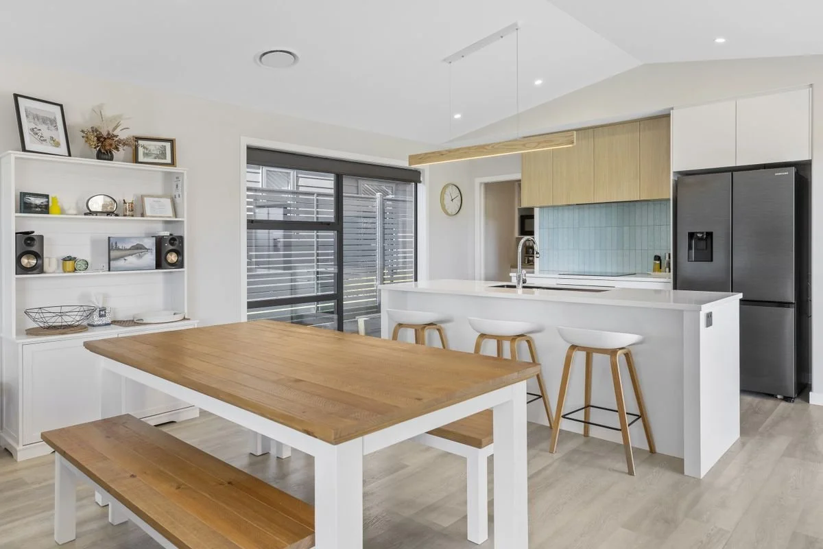Modern kitchen with white cabinetry, a kitchen island with white countertop, three white bar stools with wooden legs, a black refrigerator, a built-in microwave, and a large wooden dining table with matching benches. There is a large window and an op