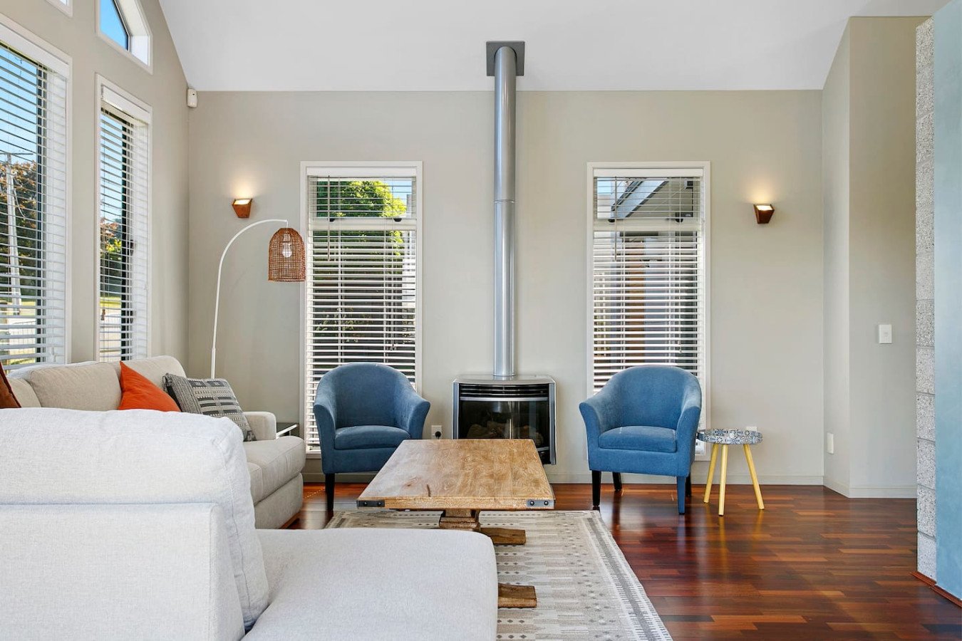 Living room with beige sofa, two blue armchairs, a wooden coffee table, a wood stove, and large windows with white blinds.