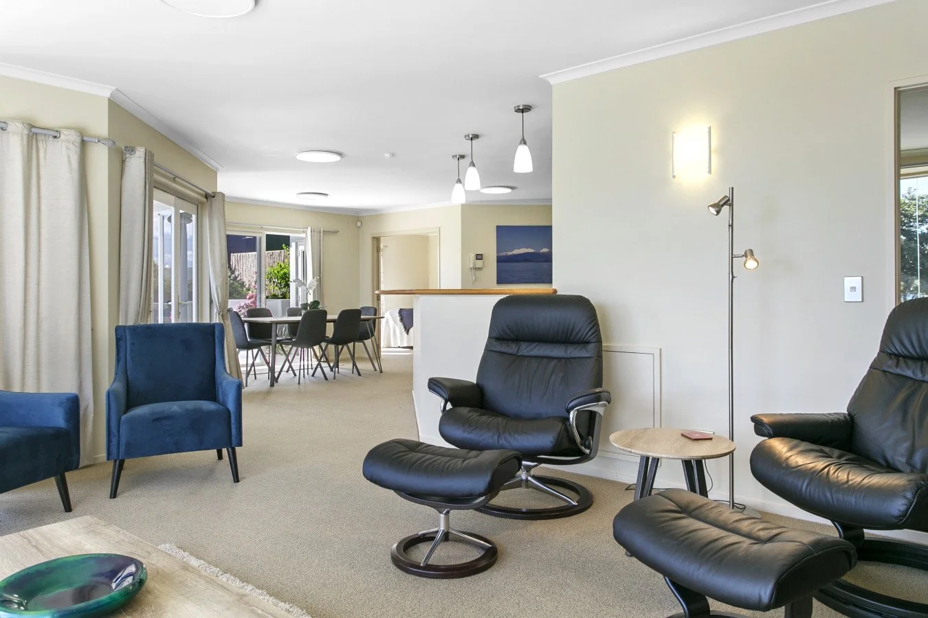Living room with black leather armchairs and a blue armchair, small side table, white curtains, windows and sliding doors leading to a garden, dining area with a round table and six chairs, beige walls, ceiling lights, and a lamp.