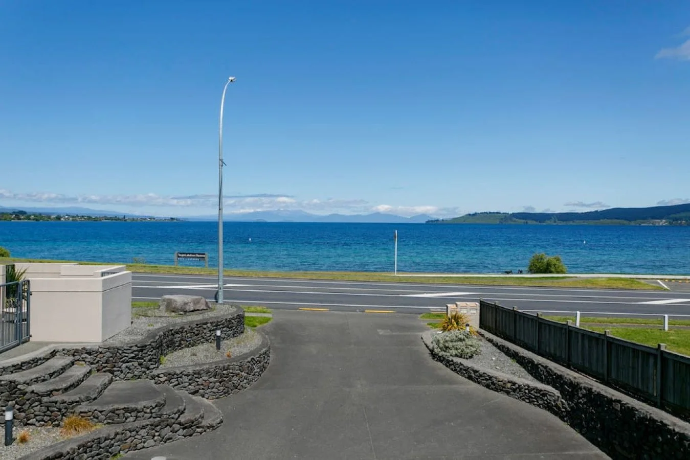 View of a body of water, possibly a lake, with hills in the background, a clear blue sky, and a parking area in the foreground with a stone and concrete steps, a streetlamp, and a landscaped area.