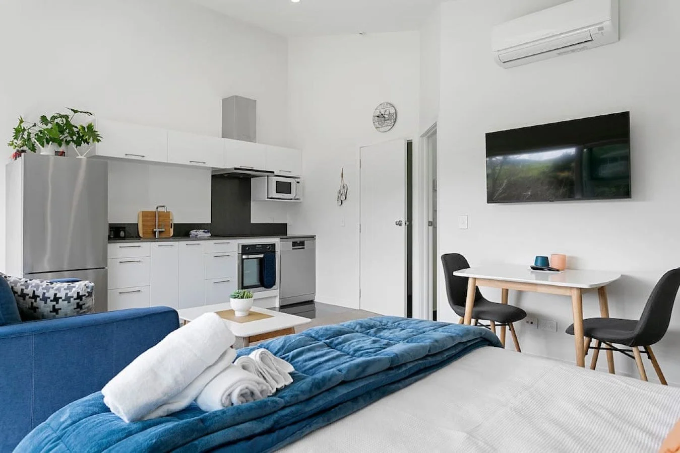 A studio apartment with a kitchen, a bed, a small dining table with two chairs, a television on the wall, and an air conditioner.