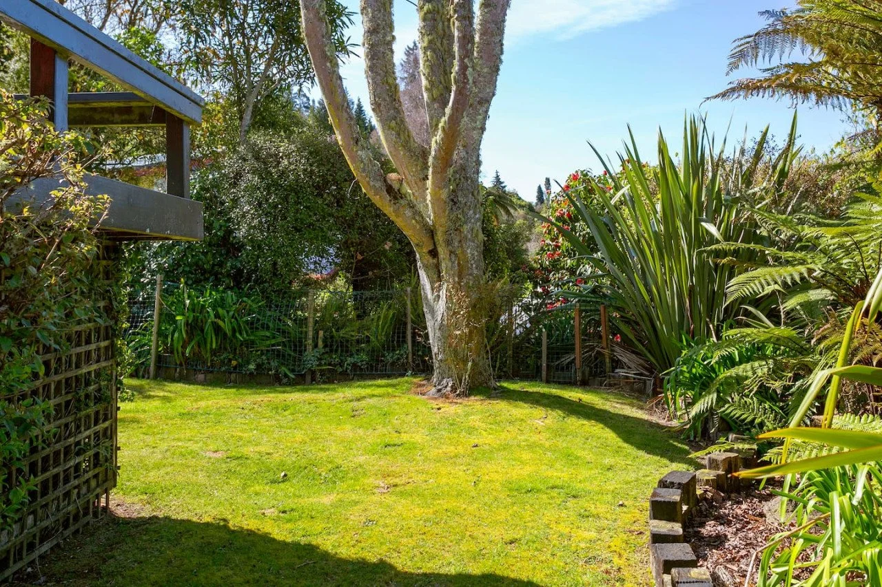 A backyard garden with a large tree at the center, surrounded by lush green plants and shrubs, with a wooden deck on the left and a grassy area.